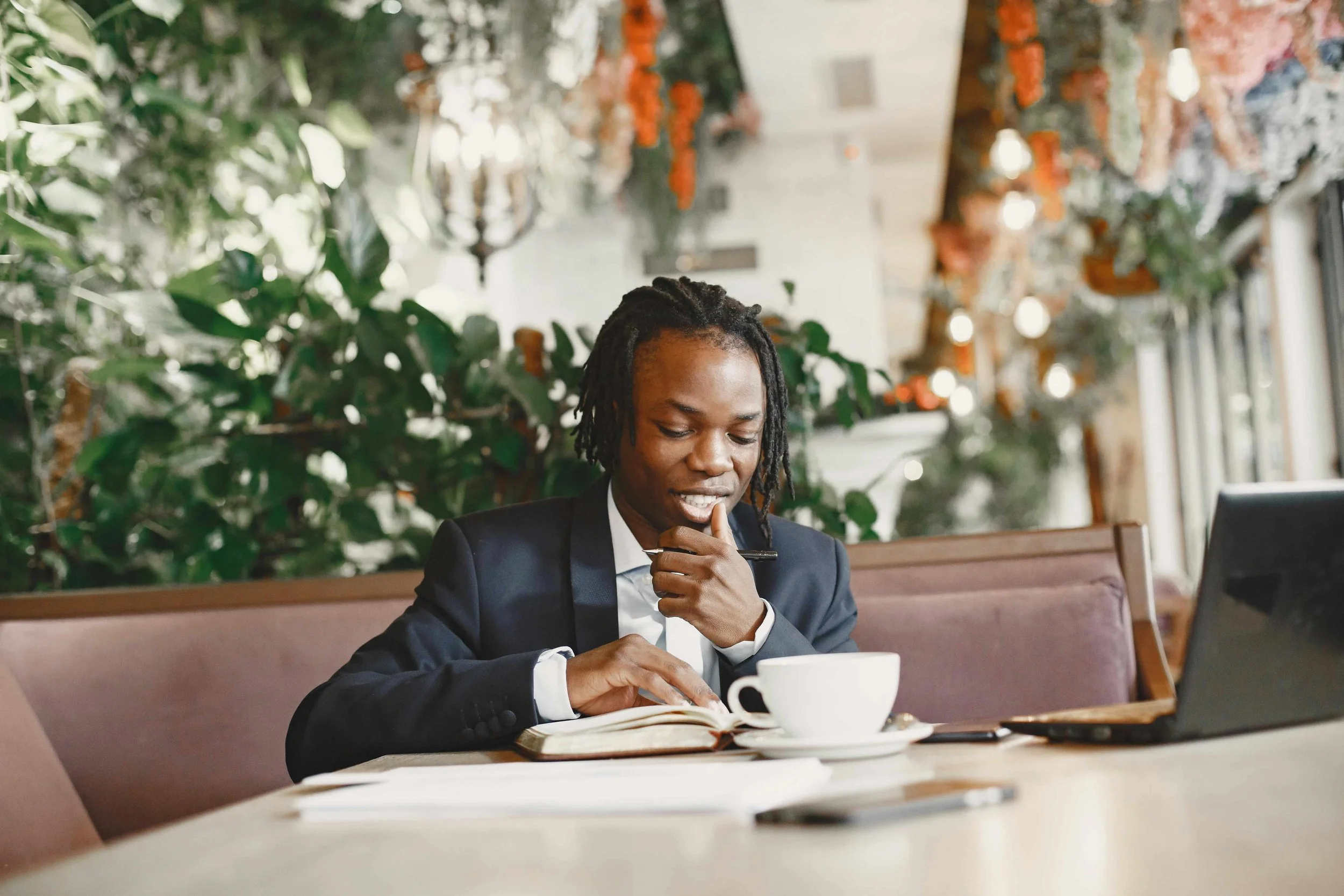 A man in a business suit sitting at a table in a cafe or restaurant, reading a book and holding a pen, with a cup of coffee in front of him, and greenery and decorative lighting in the background.