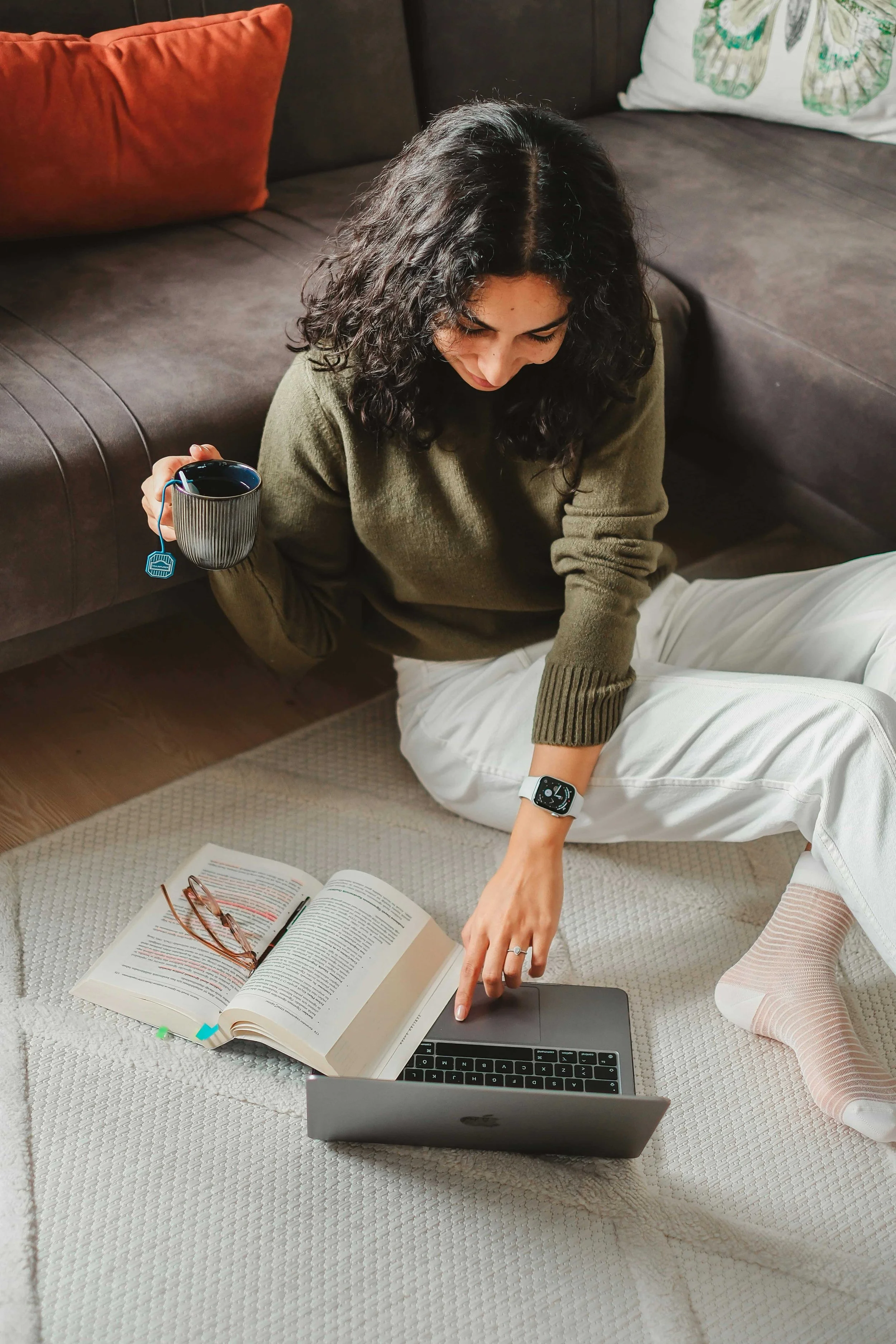A woman with curly dark hair, wearing a green sweater and white pants, sitting on a beige textured blanket on the floor, pointing at a laptop with her right hand, with an open book and reading glasses on the book, a cup of coffee in her left hand, and a smartwatch on her right wrist. Behind her is a dark gray couch with an orange pillow and a white pillow with green butterfly design.