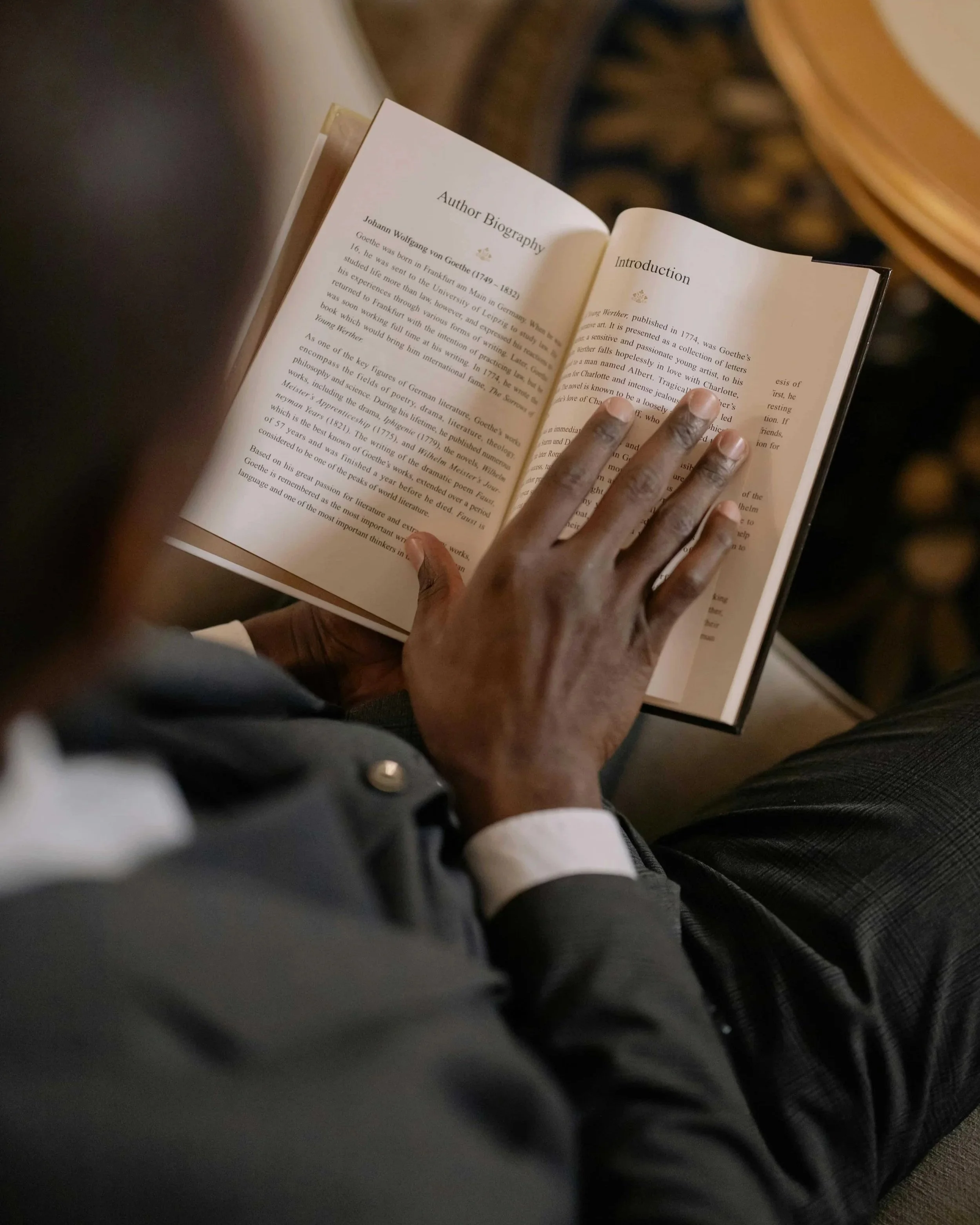 A person with dark skin and gray hair is holding an open book and reading. The person's hand is visible resting on the right page, and they are wearing a business suit with a white shirt and dark pants. The background shows part of a chair with a floral or ornate design.