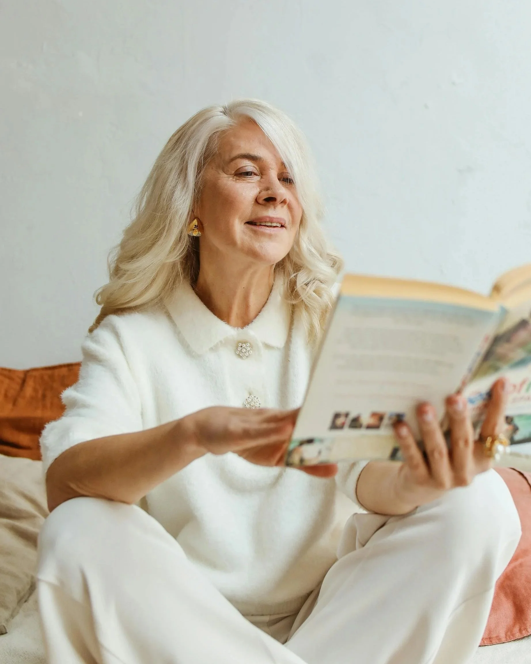 An older woman with long gray hair wearing a white coat, earrings, and a brooch, sitting on a couch and reading a magazine in a room with white walls.