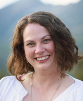 A woman with shoulder-length curly brown hair smiling outdoors with mountains in the background.