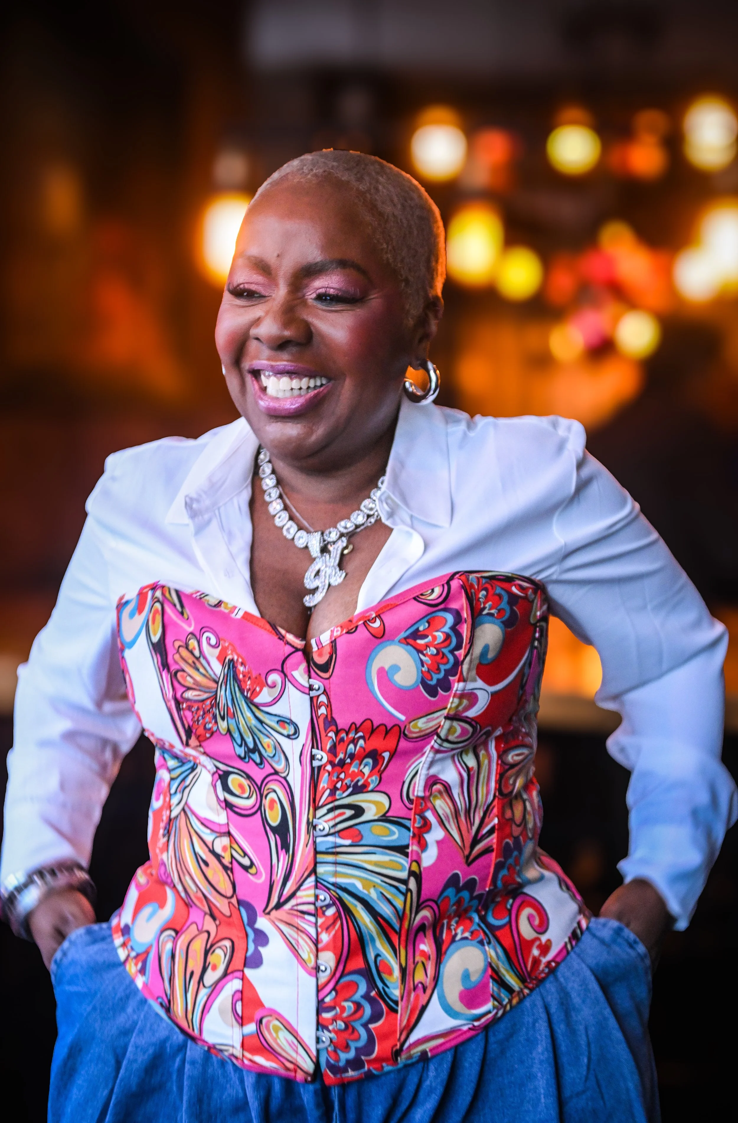 A smiling older woman with short gray hair, dressed in a white blouse and colorful, patterned corset top, standing with hands in her pockets in a warmly lit indoor setting.