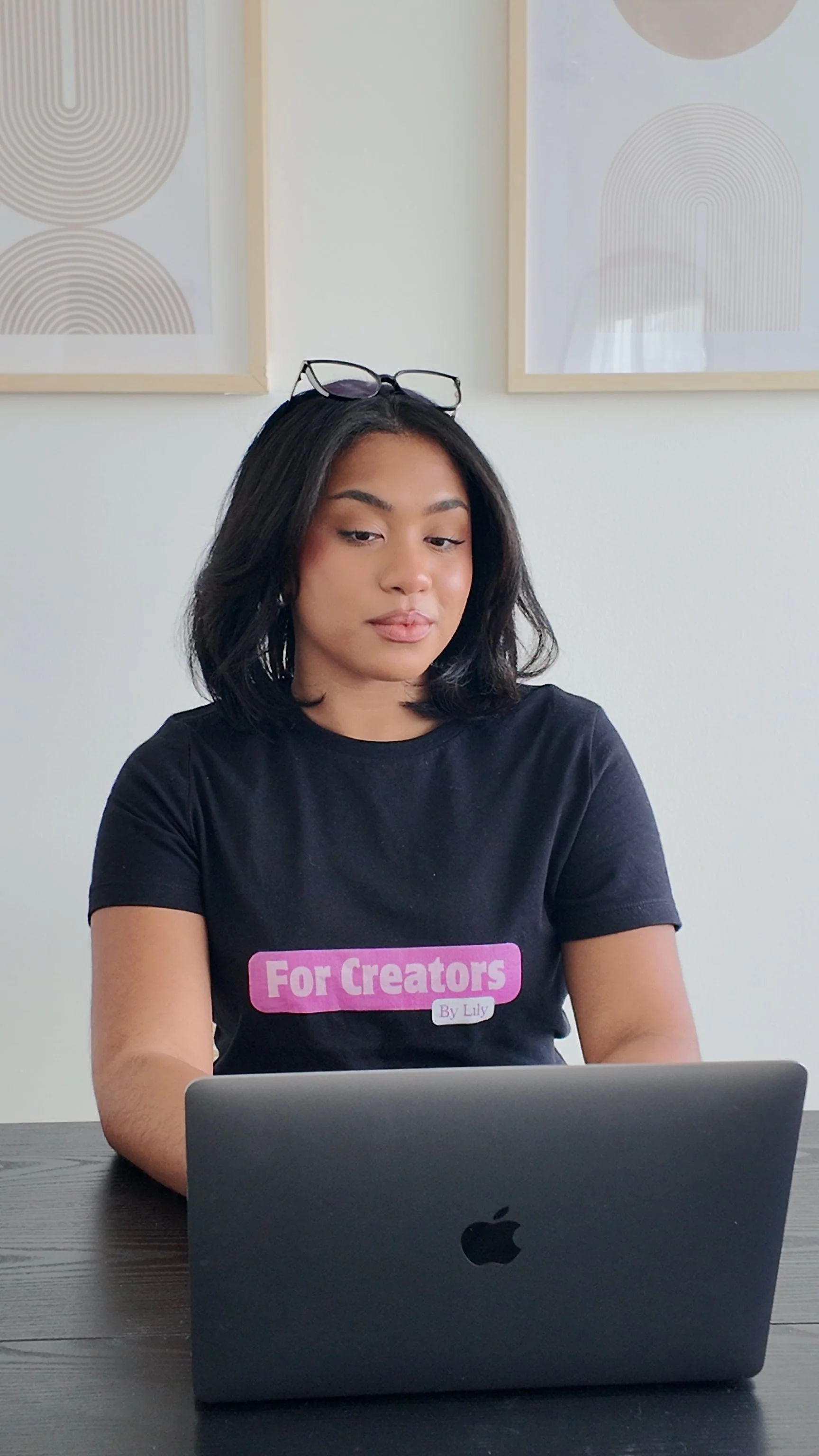 A woman with shoulder-length black hair, wearing a black T-shirt that says "For Creators By Lily," is sitting at a dark wooden table using a closed silver MacBook laptop. She has black-framed glasses resting on her head, and is looking at her laptop. The background features two framed abstract art prints on a white wall.