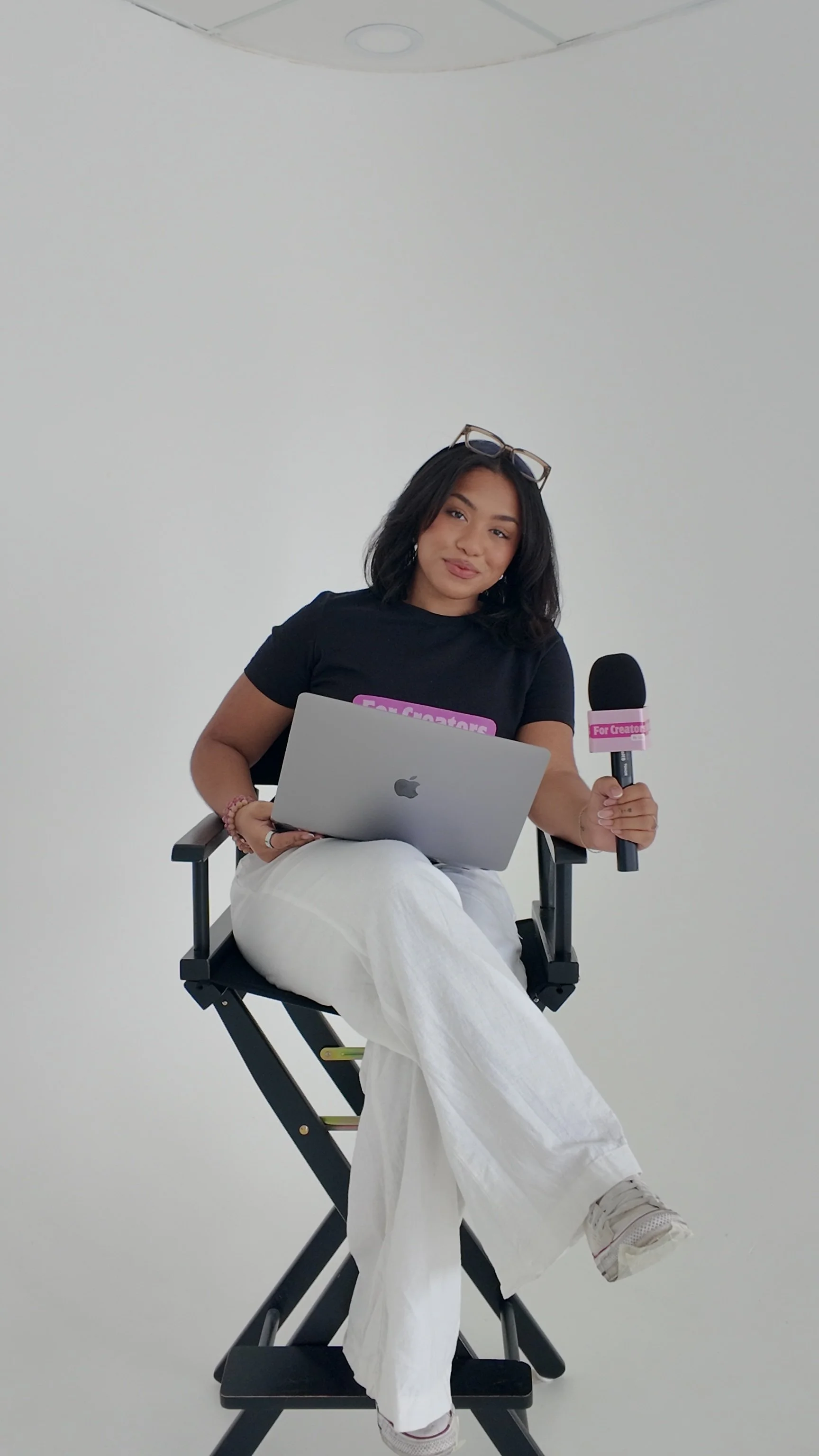 A woman in a black t-shirt and white pants sitting on a director's chair, holding a laptop and a microphone, with glasses on her head, against a plain white background.