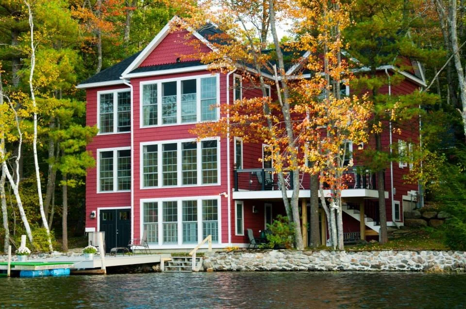 Lake side of the red three-story lakehouse with a deck, stone shoreline wall, and dock