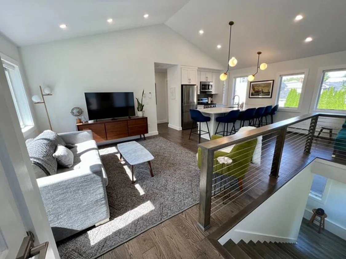 Open-concept living room and kitchen with a gray sofa, a wooden TV stand, a flat-screen TV, and a small marble coffee table. The kitchen has a white island with blue barstools, a stainless steel refrigerator, and modern lighting fixtures. Large windows let in natural light, and there's a wooden railing beside a staircase.