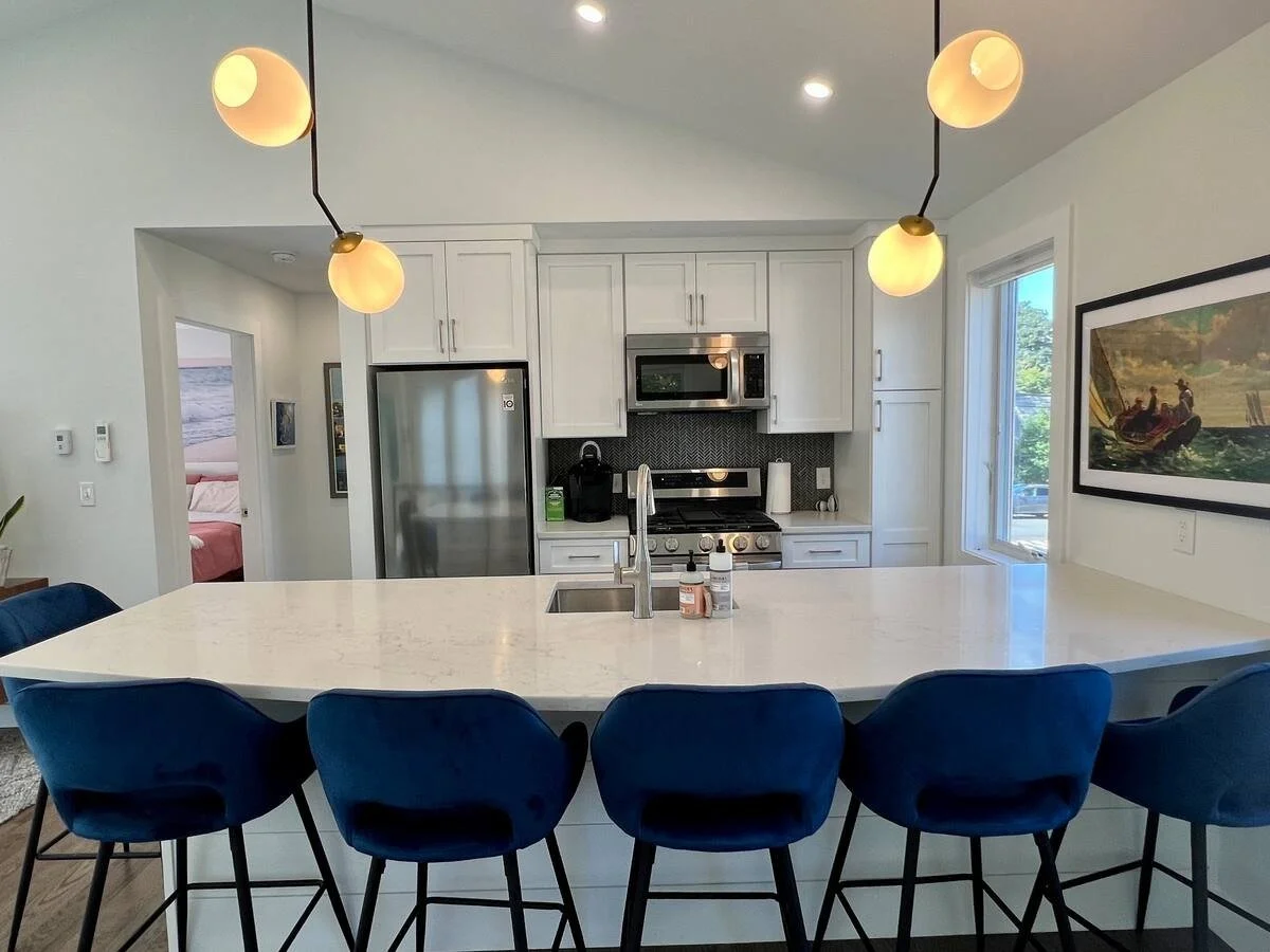 Modern kitchen with white cabinets, stainless steel refrigerator, microwave, and stove. A white marble island with blue chairs, pendant lights, and a framed painting on the wall.