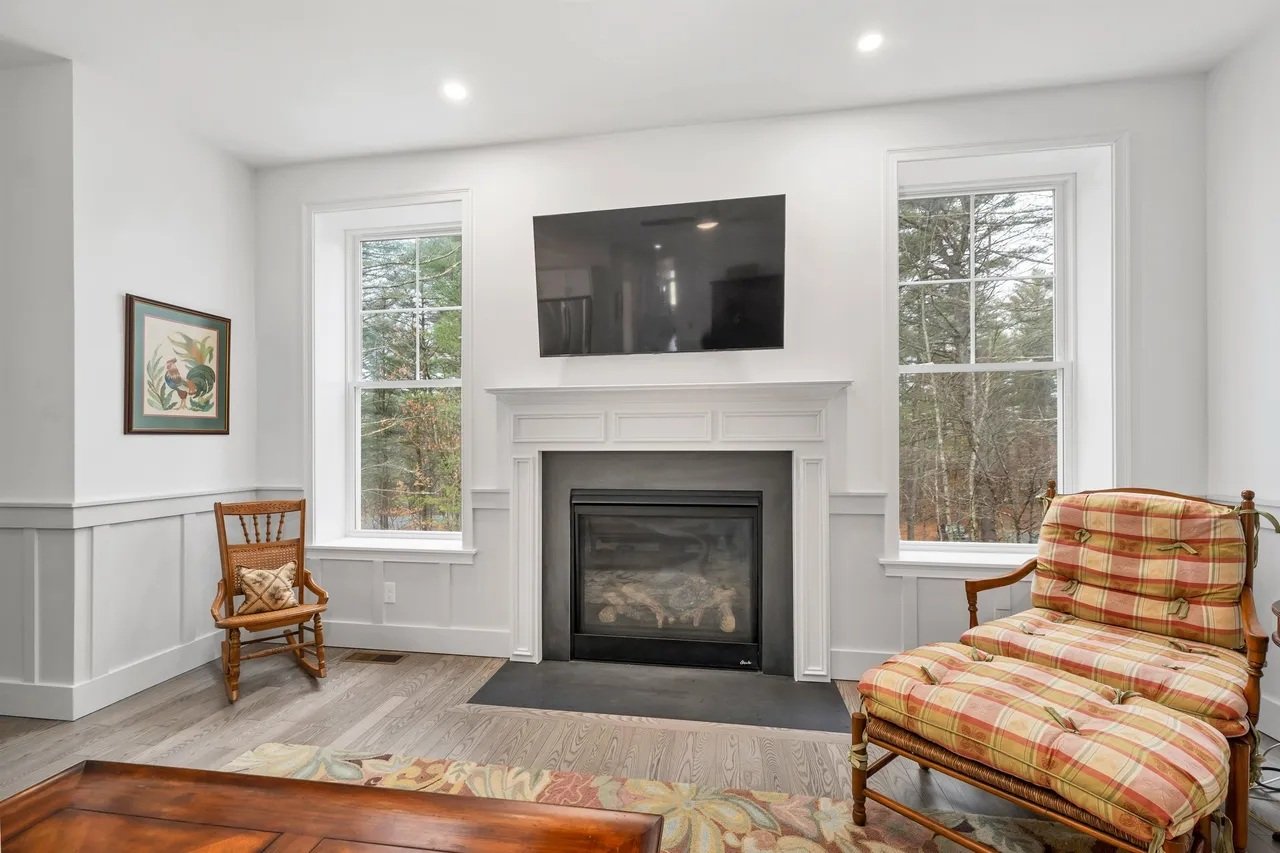 Living room with a white fireplace, two large windows, a wall-mounted black TV above the fireplace, a wooden chair with a cushion, a plaid cushioned armchair, and a wooden coffee table.