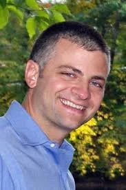 Smiling construction professional outdoors wearing a blue collared shirt, standing in front of green foliage.