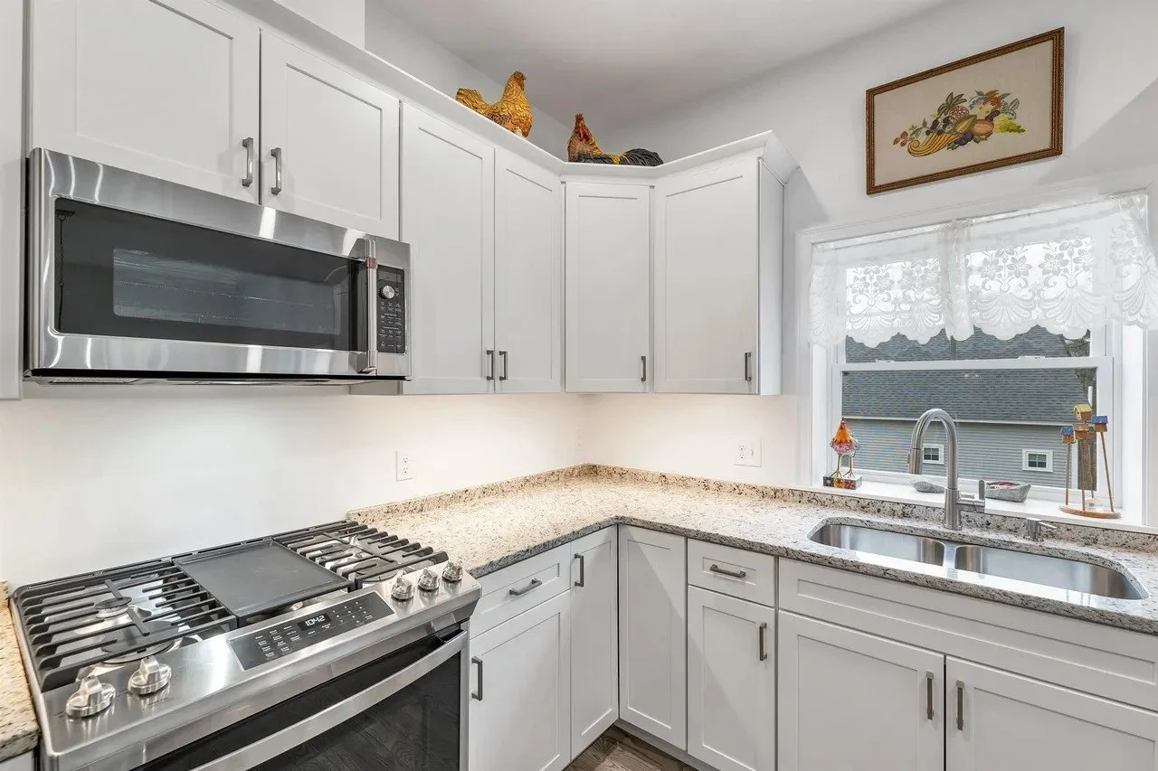 Kitchen with white cabinets, granite countertops, stainless steel microwave and stove, window with lace curtains, decorative chicken figurines on cabinet, framed artwork above window, and a double sink.