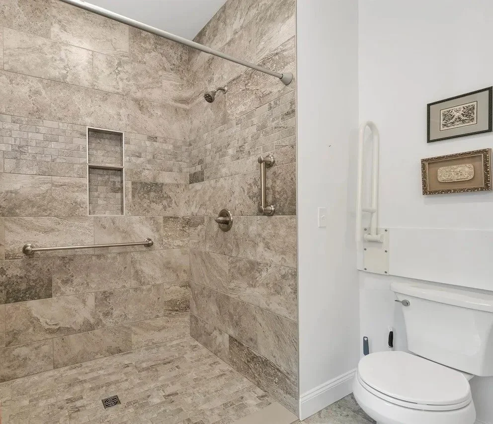 Bathroom with a walk-in shower and toilet. The shower has beige and gray stone tiles, a built-in niche, and metal grab bars. The toilet area has framed artwork on the white wall.