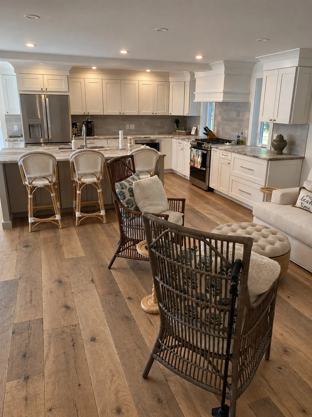 White kitchen with a gray-brown island, rattan bar stools, plaster hood, and wicker lounge chairs on oak floors