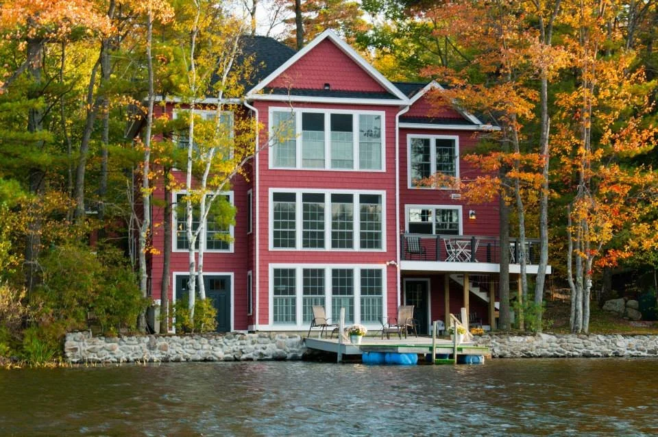 Red-sided traditional lakehouse with large windows and a dock on the shoreline, surrounded by autumn foliage
