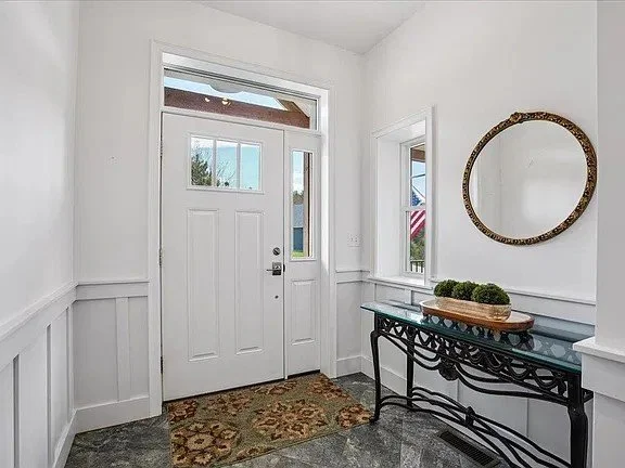 Foyer with a white front door, wainscot paneling, a round mirror, and a console table