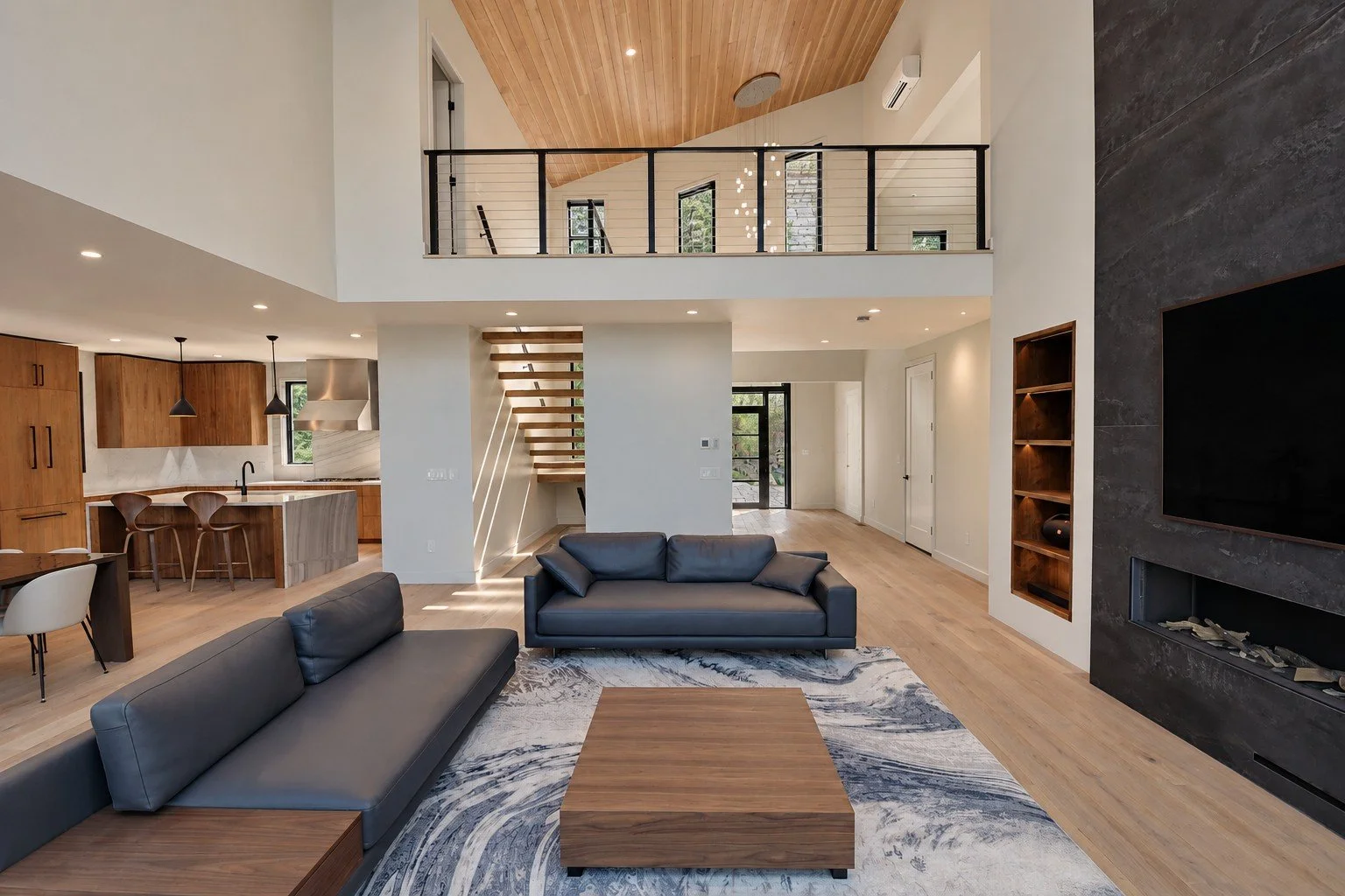 Open living room with a vaulted ceiling, a dark stone fireplace wall, gray sofas, and the kitchen beyond