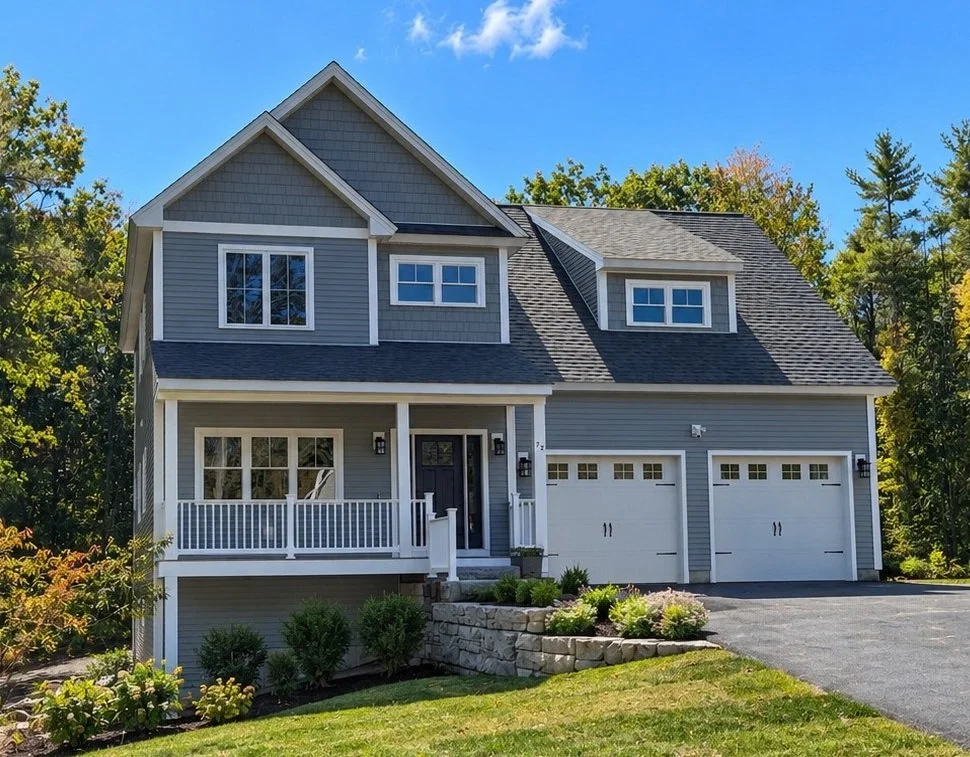 Front of a gray two-story spec house with a covered porch, two-car garage, and stone retaining wall