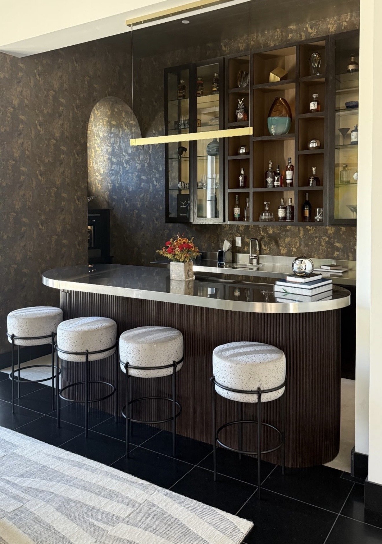 Modern kitchen with a black and brown color scheme, a curved dark wood bar with a silver countertop, four white speckled barstools with black metal frames, a black textured wall, and open dark wood shelves with bottles and glassware.