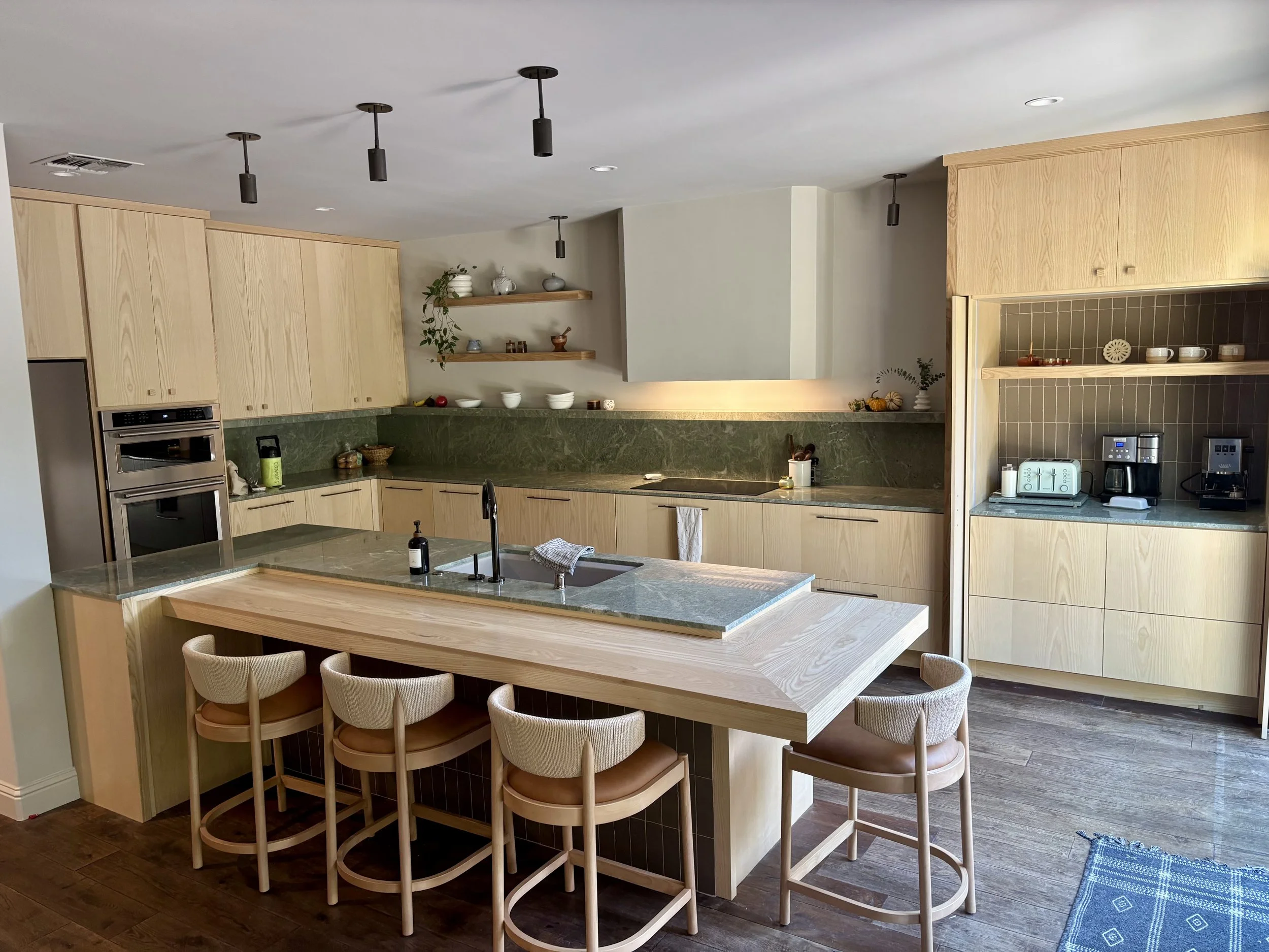 Modern kitchen with light wood cabinetry, green marble countertops, and a kitchen island with seating for five. The kitchen features built-in ovens, a sink with a black faucet, and various appliances including a toaster, coffee maker, and kettle. Open shelves hold dishes, bowls, and decorative items. A small towel hangs from the oven handle, and a soap dispenser is on the island.