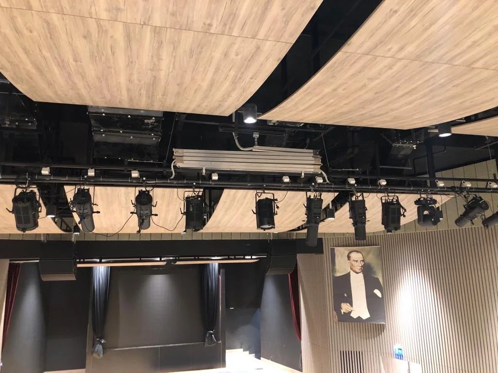 A stage in a theater or auditorium with black curtains, wood paneling on the ceiling, and stage lighting equipment hanging from the ceiling.