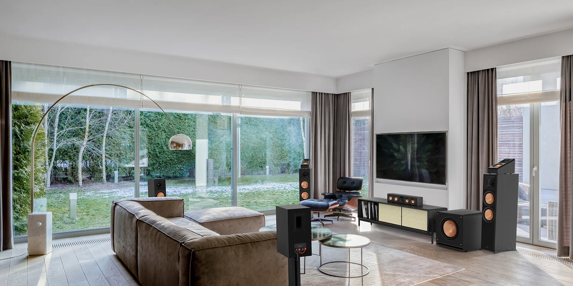 Modern living room with large glass sliding doors, beige sofa, black leather lounge chair, flat-screen TV, and high-end speakers.