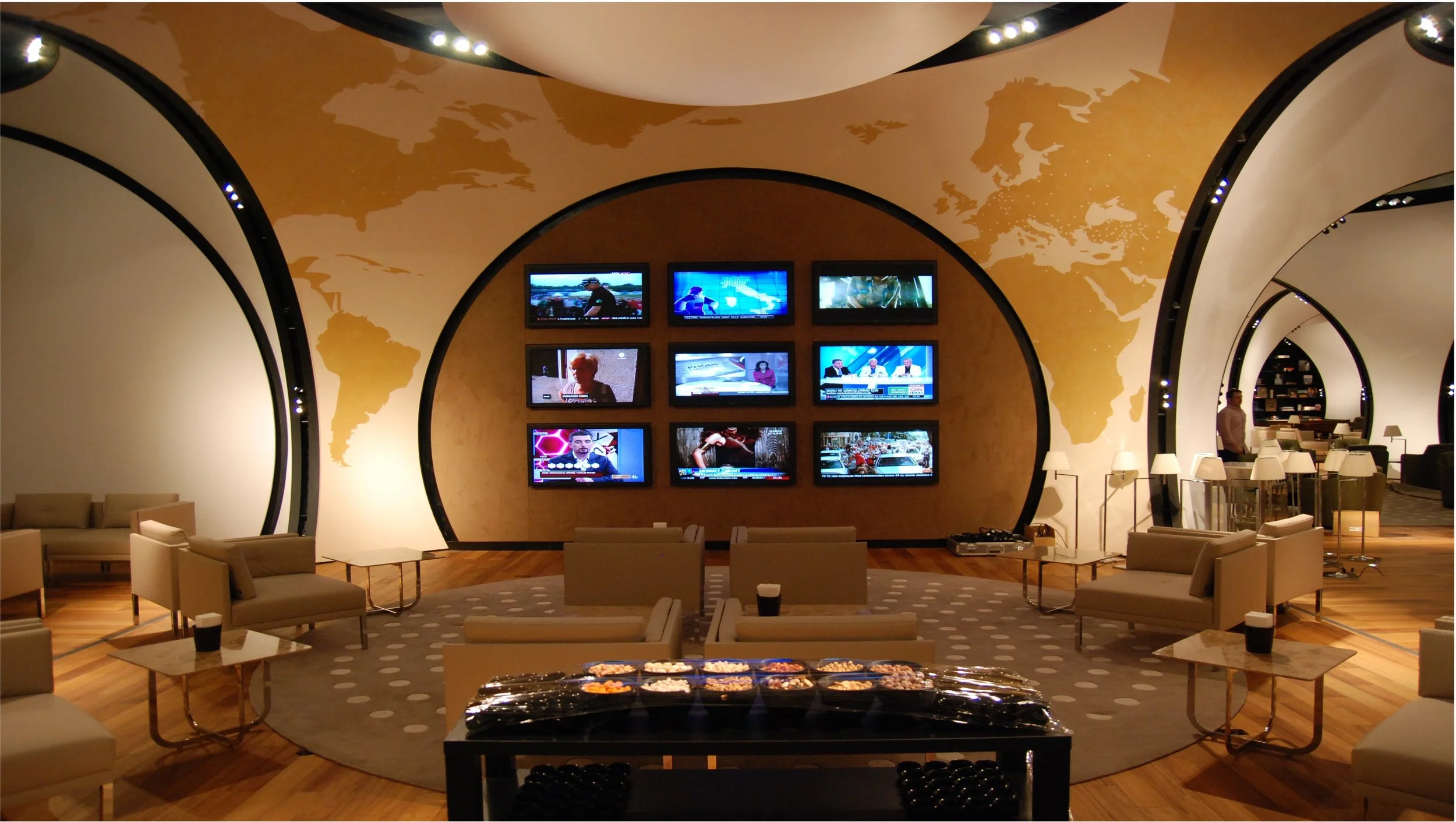 Modern lounge area with beige chairs and tables, a circular patterned rug, and a wall decorated with a gold world map. Multiple flat-screen TVs display news and sports channels. TVs have wireless headphone audio system connected.