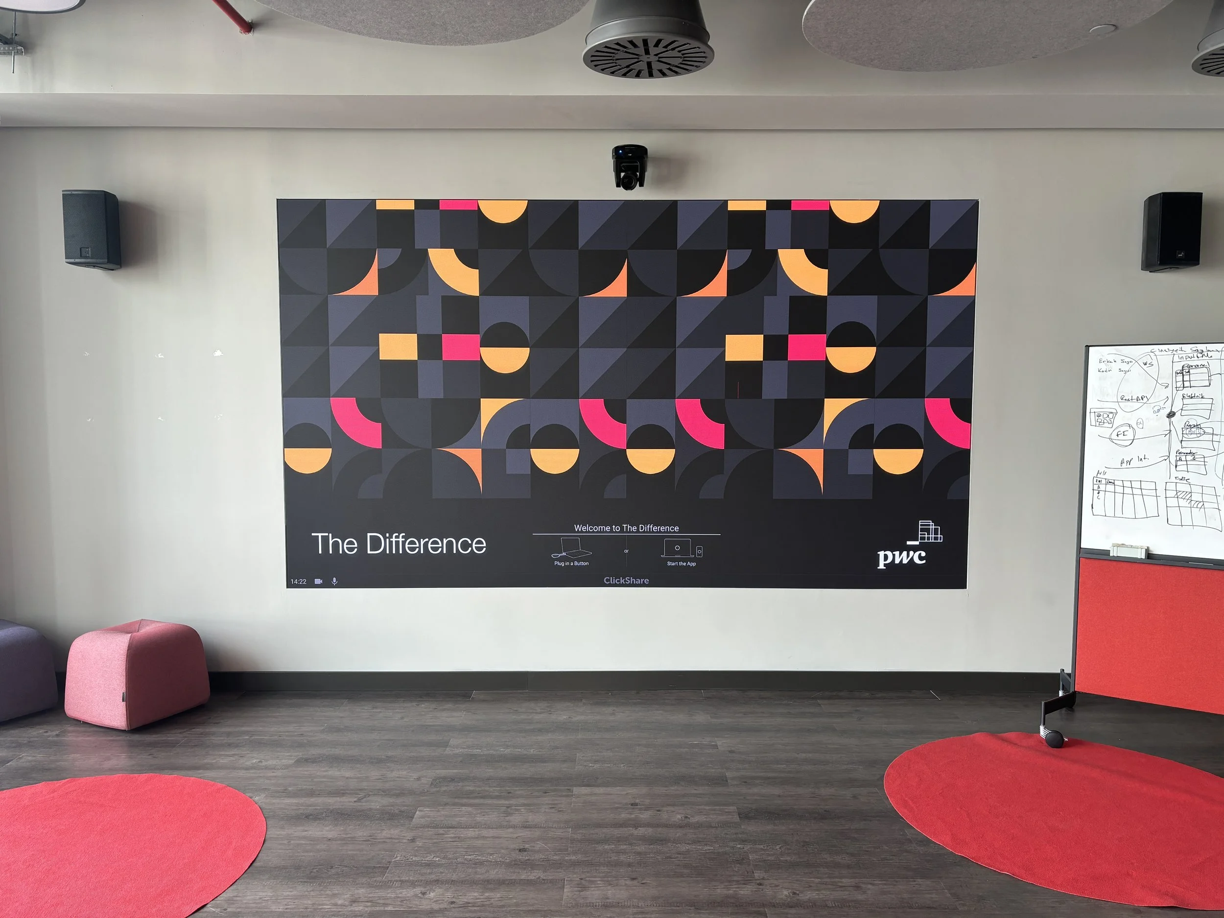 Large presentation screen displaying an abstract geometric design with black, dark gray, orange, and pink shapes, titled 'The Difference' and featuring the PwC logo, mounted on a wall in a modern conference room with two pink circular rugs and a whit