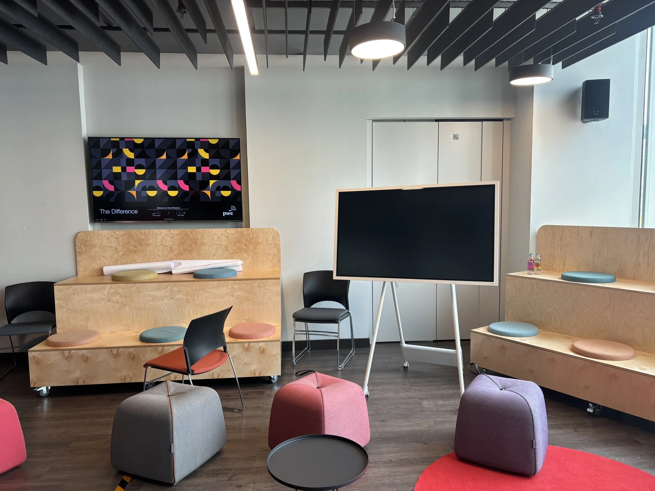 Modern conference or meeting room with chairs, colorful cushions, a large monitor, a blackboard, and a wall-mounted screen display.
