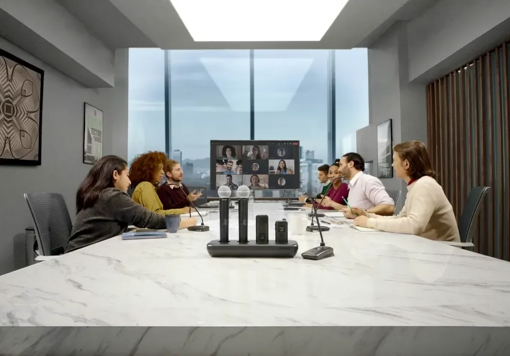 Business meeting room with eight people participating in a video conference, seated around a white marble conference table, with a large window showing a cityscape in the background.