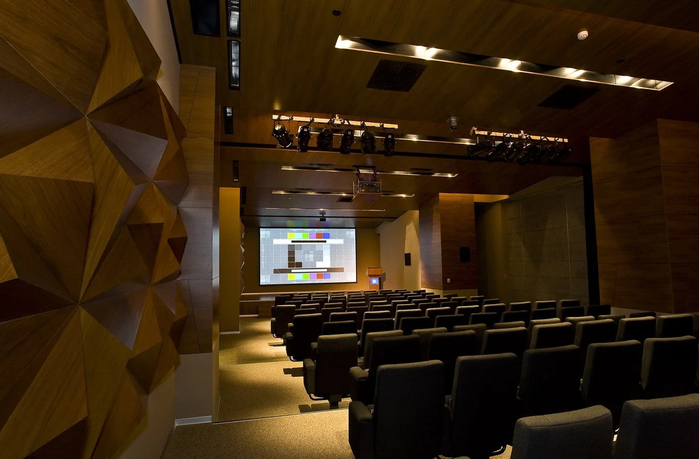 A conference room or auditorium with rows of chairs facing a large screen on the front wall. Stage lighting equipment hanging from above. There is a projector and audio video and automation systems integrated.