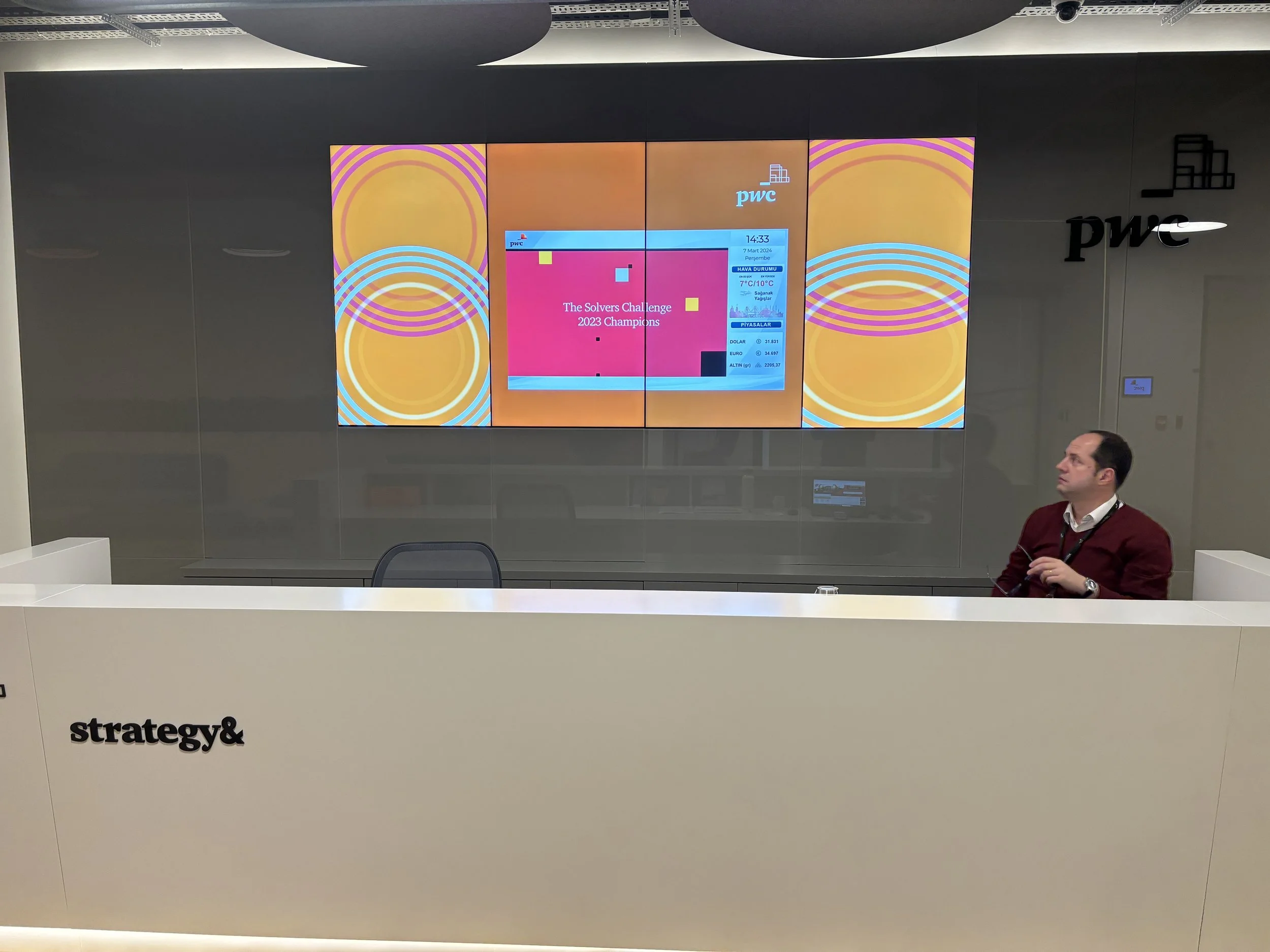 A man in a red sweater sitting at a white reception desk with the word 'strategy&' on it, in front of a large display screen showing colorful graphics, the PwC logo, and various data including the time 14:33 and date March 7, 2024.