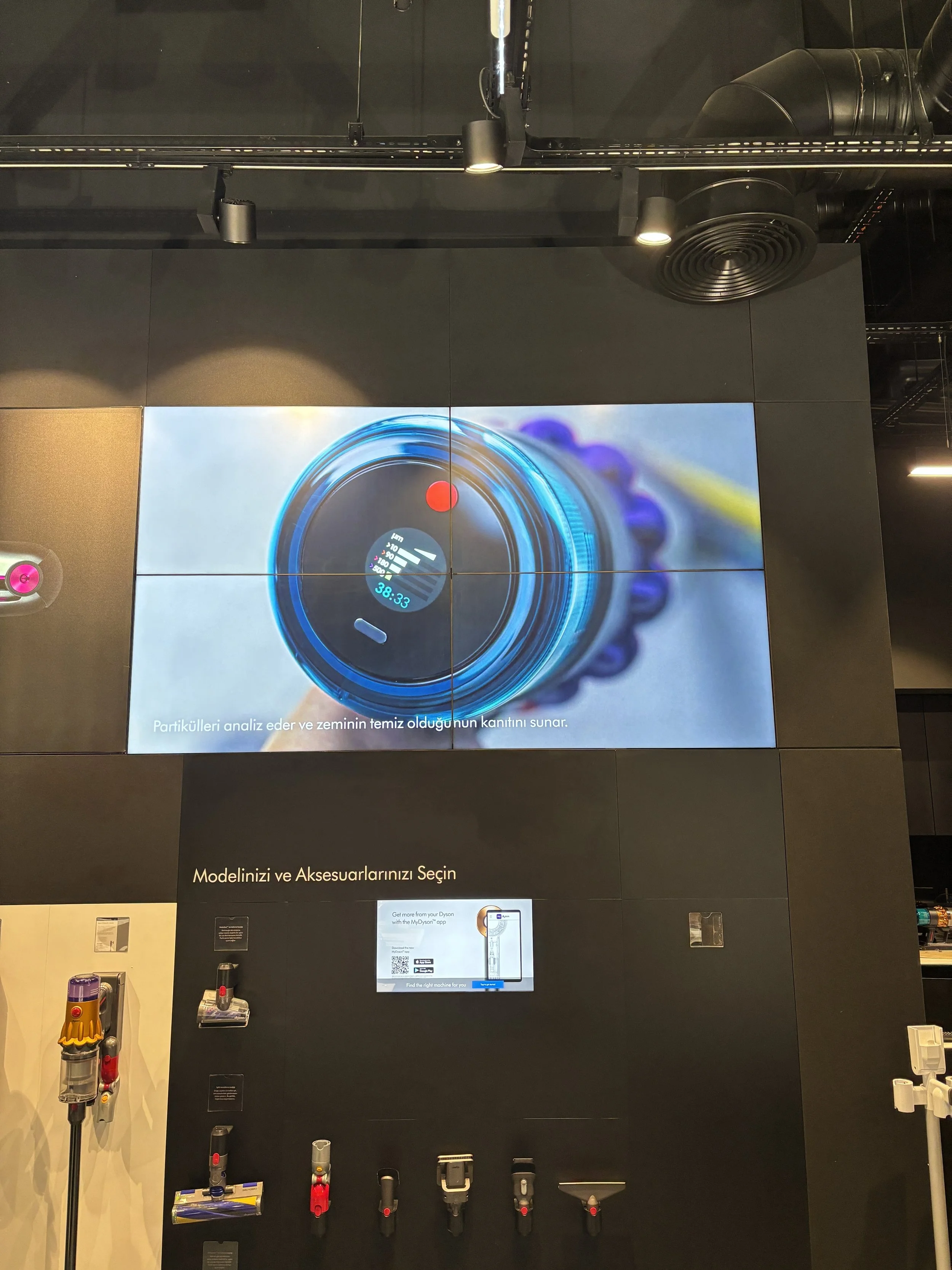 Large digital screen displaying a close-up image of a vacuum cleaner filter, with a red point and a digital reading of 38.33. Below the screen, there are various vacuum cleaner attachments mounted on a black wall, with a smaller screen and a QR code 