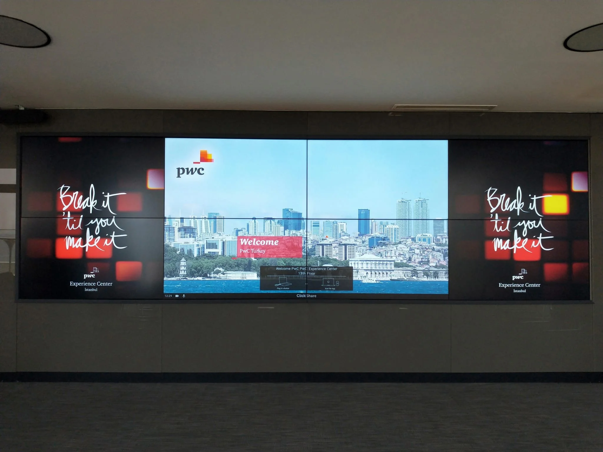 Large digital display wall showing a cityscape with high-rise buildings, a blue sky, and water in the foreground. The display features branding for PwC at an experience center.