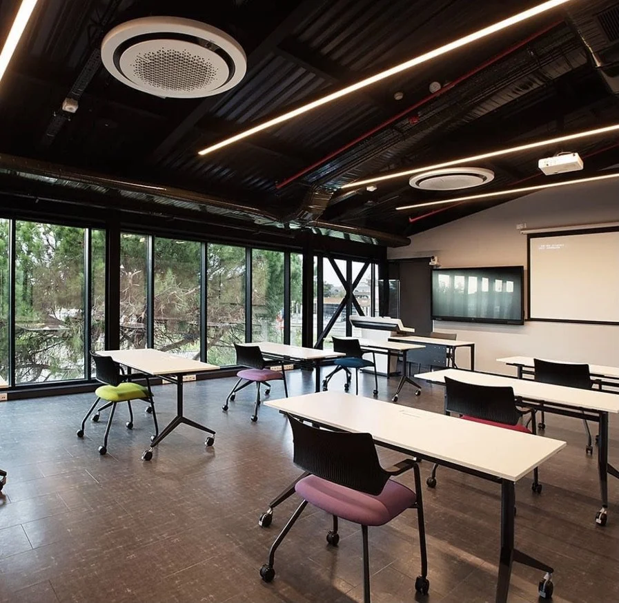 Modern classroom or meeting room with white tables, black chairs with colorful cushions, large floor-to-ceiling windows, and a projector screen. The room has a dark ceiling with circular light fixtures and linear LED lighting.