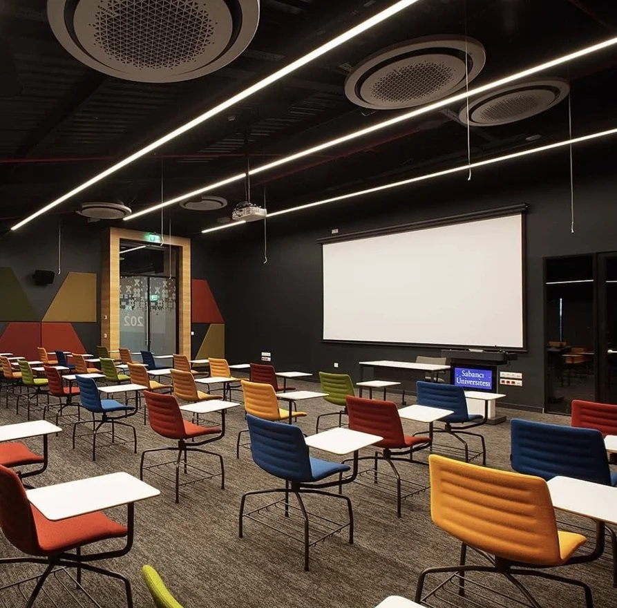 An empty classroom or conference room with colorful chairs and white desks facing a large white projection screen on a black wall, with modern ceiling lights and acoustic panels. This is a hybrid university auditorium.