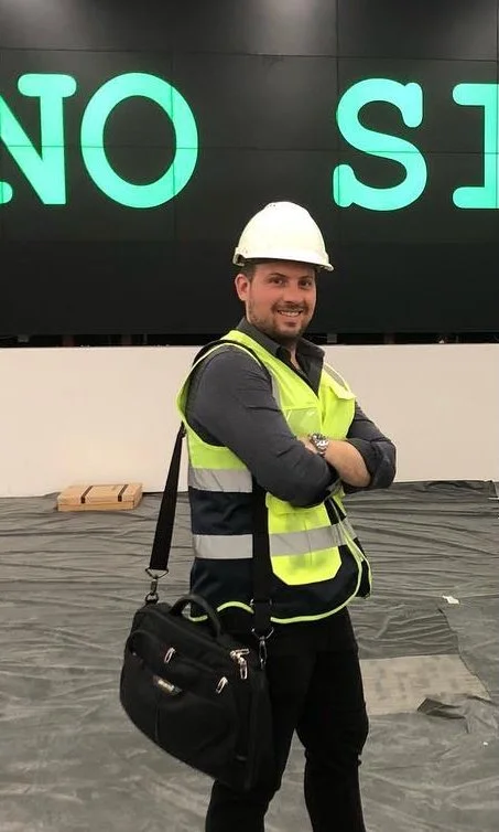 Man wearing a white safety helmet and a yellow safety vest standing with arms crossed in front of a large digital display board. The man is smiling and carrying a black shoulder bag.