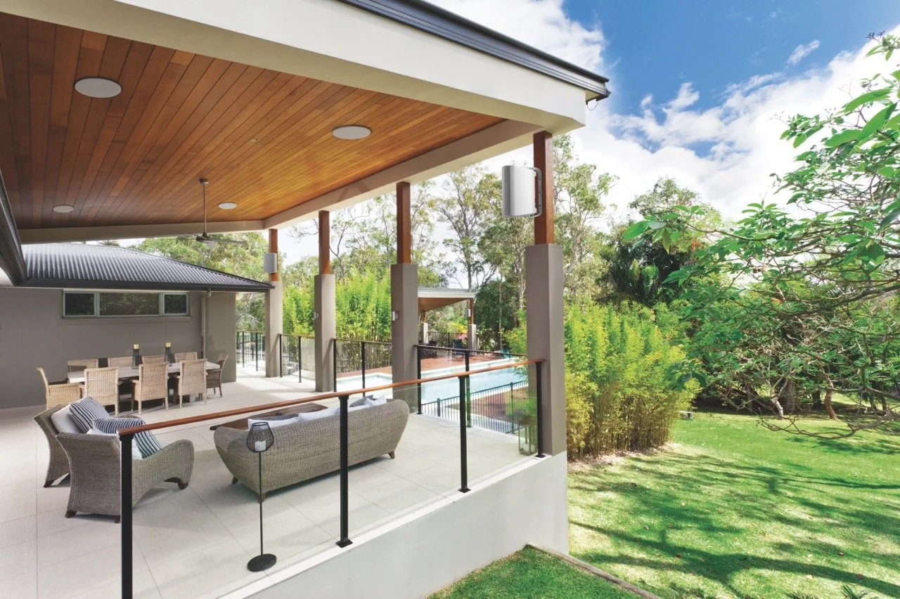 A covered outdoor patio with wicker furniture, a dining table, and lush green trees in the backyard. There is an outdoor audio sound system installed.