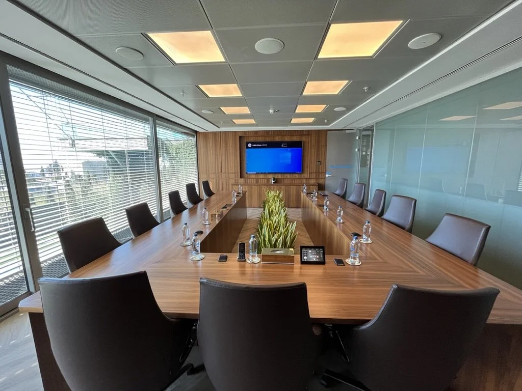 Modern conference room with a long wooden table, black chairs, a large screen at the front, and a skylight ceiling.