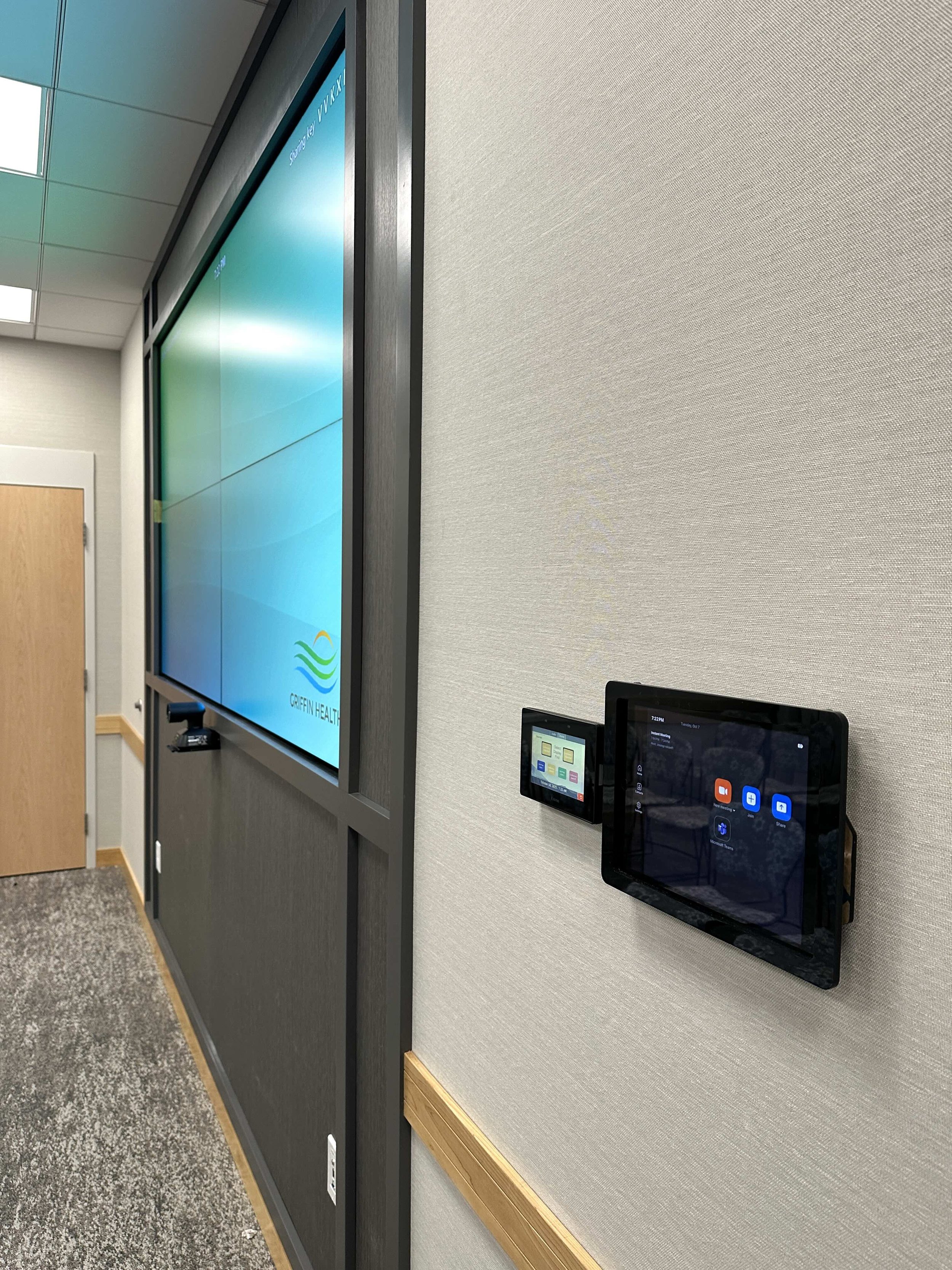 A wall with a large flat screen monitor displaying a logo and text, alongside two smaller control panels with touchscreens, in a modern office or conference room.