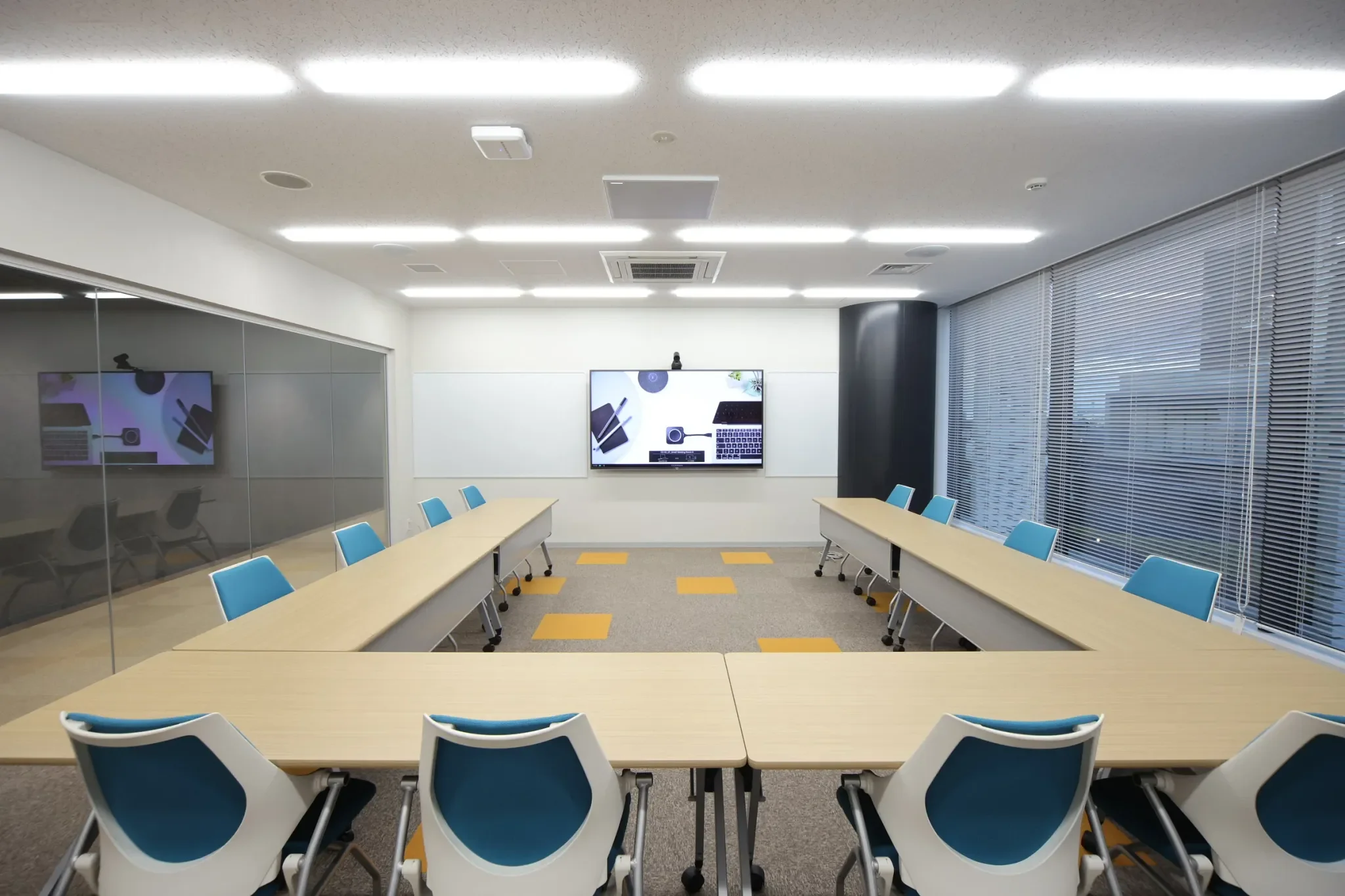 Empty modern conference room with U-shaped tables, blue chairs, a large screen on the white wall, and window blinds