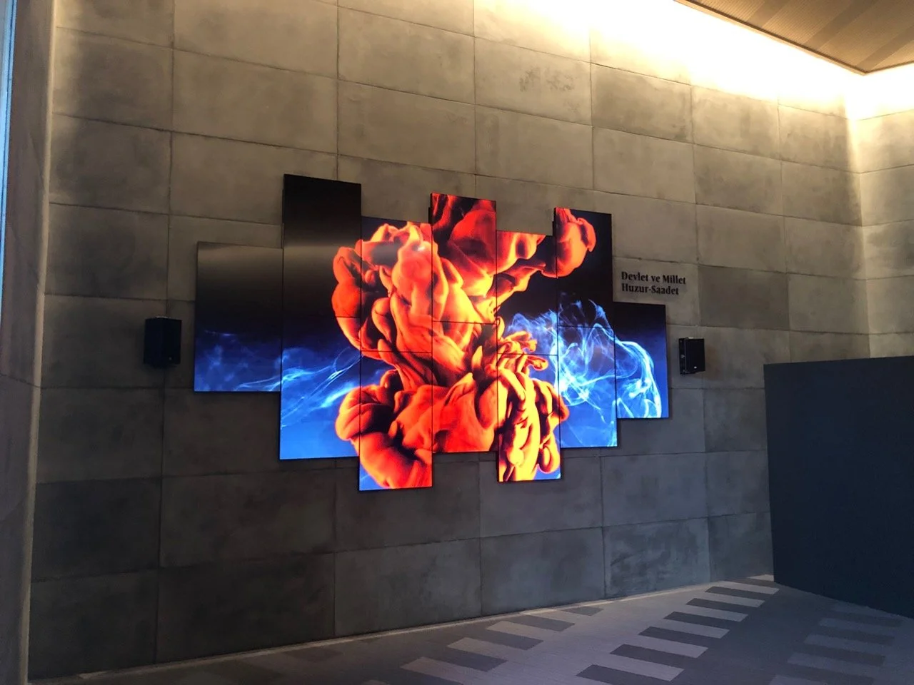 A multi LCD monitor digital art installation on a gray stone wall showing an abstract explosion of orange and blue ink or smoke.