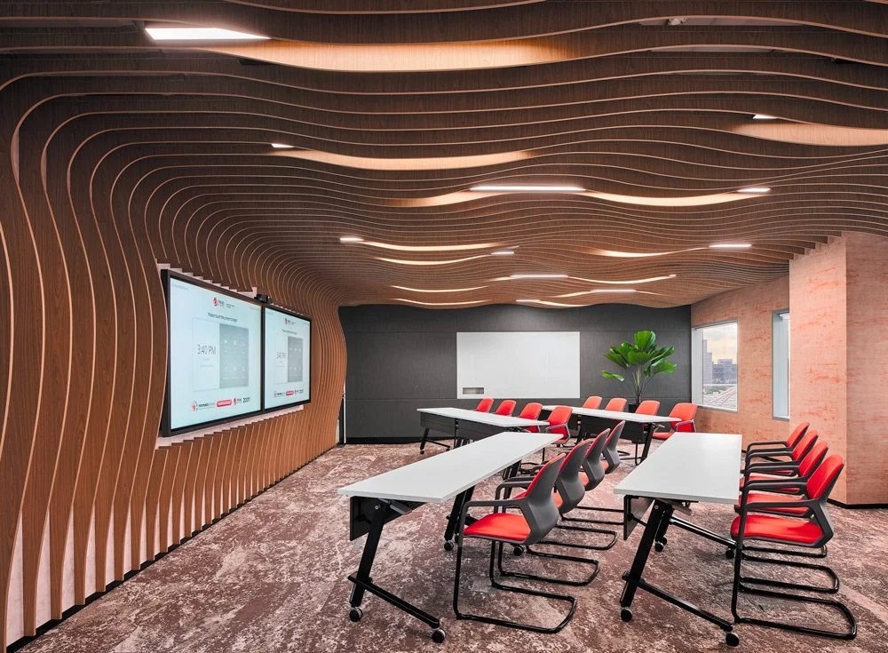 Modern conference room with wavy wooden ceiling, carpeted floor, multiple flat-screen monitors, red and black chairs, white tables, and a potted plant in the corner.