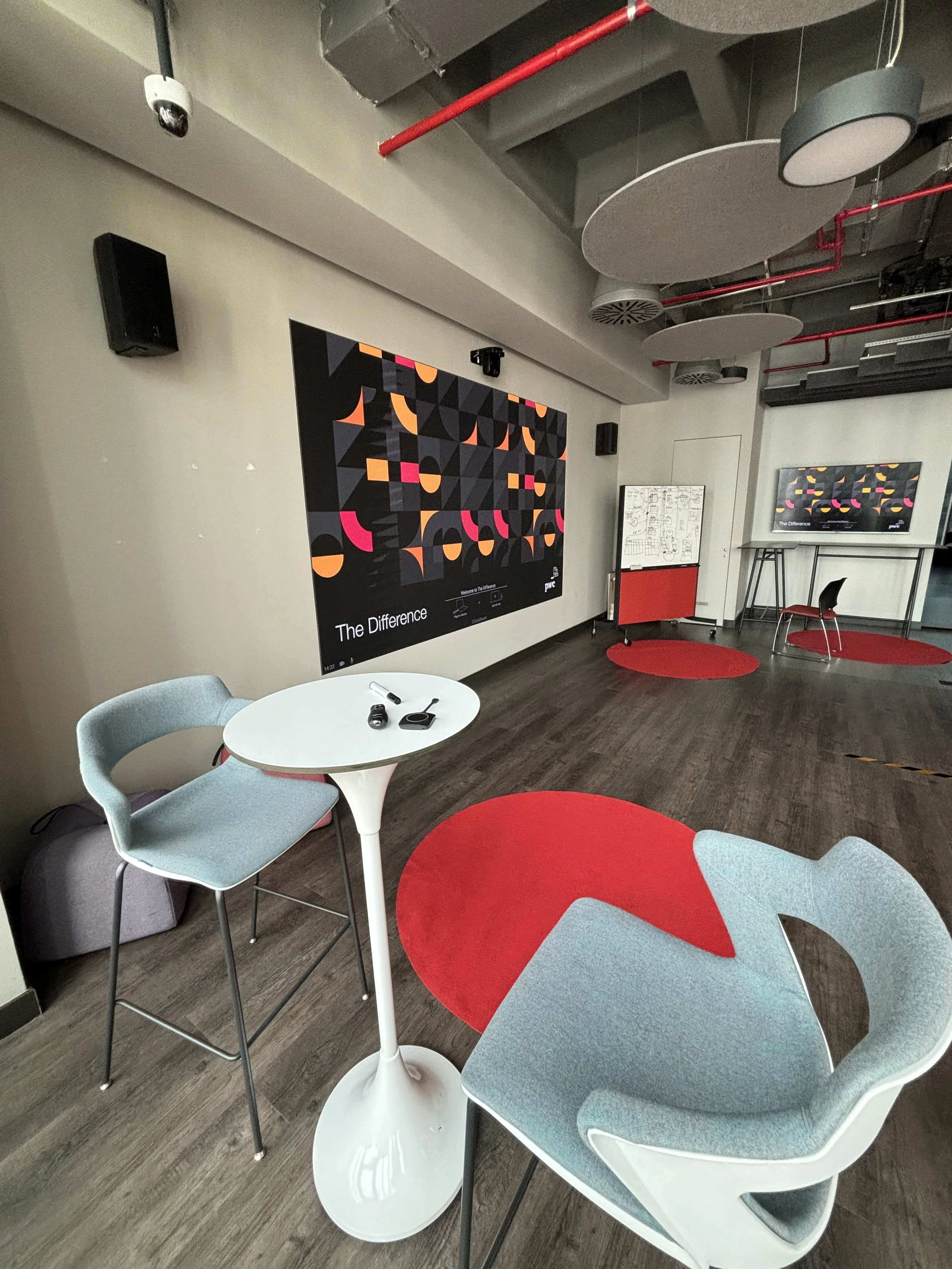 An interior room set up for a presentation or discussion, with a large wall display titled 'The Difference', two chairs around a tall white table, and a television screen displaying the same graphic as the wall. The room has red circular rugs, ceilin