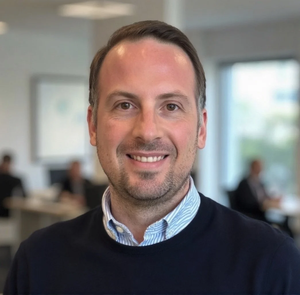 A professional headshot of a man with short brown hair, a trimmed beard, wearing a light blue striped dress shirt and a black sweater, smiling in an office setting with blurred background of colleagues and large windows.