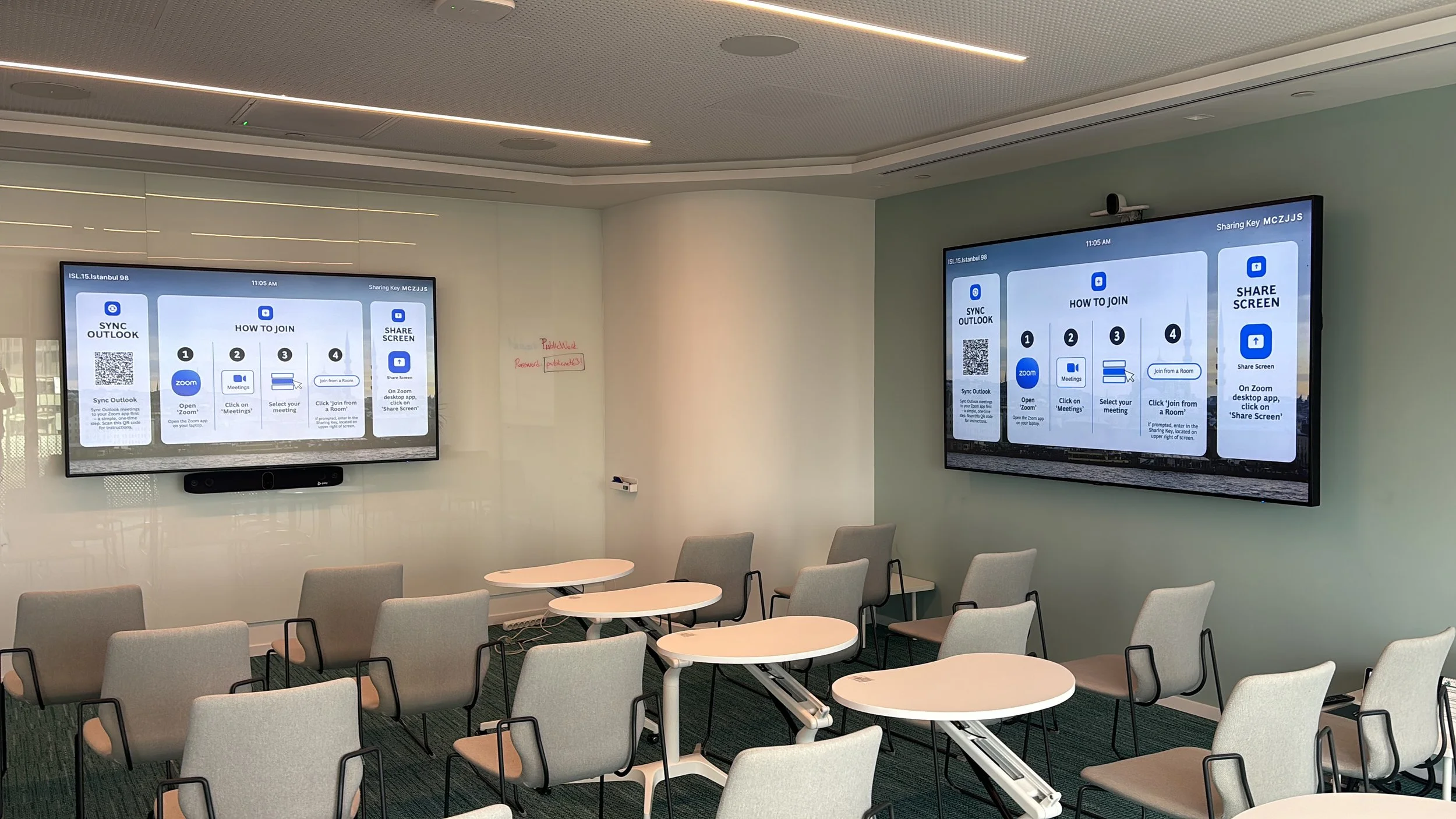 A conference room with multiple beige chairs and white circular tables arranged in rows. Two large digital screens on the wall display instructions for joining a meeting via Zoom.