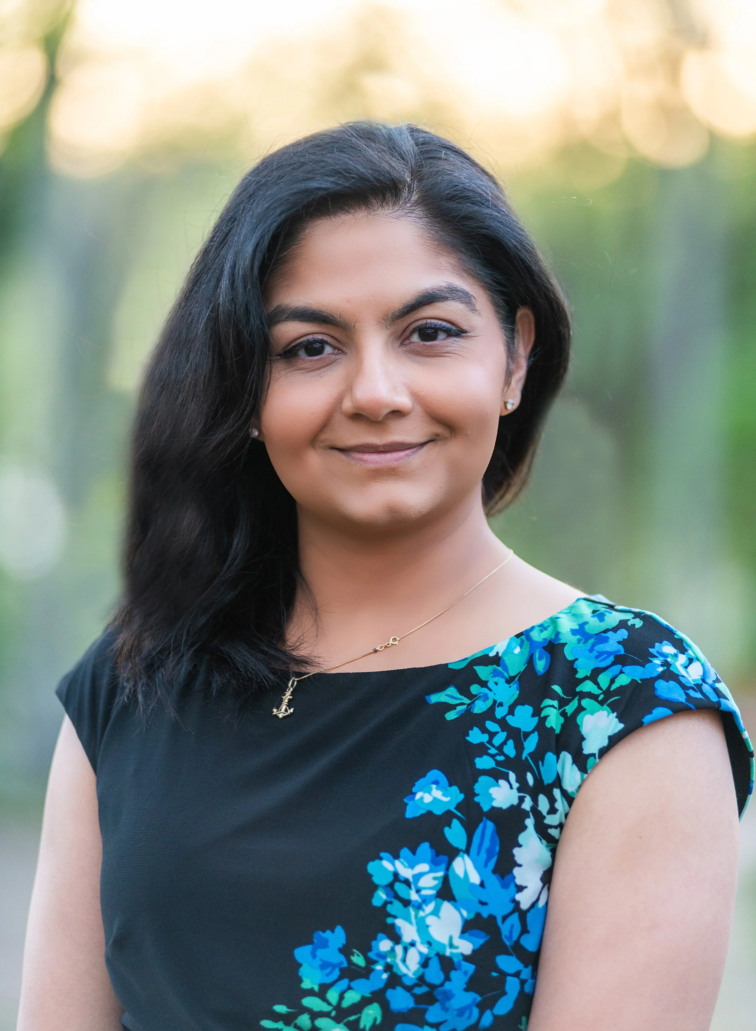 A woman with dark, wavy hair smiling outdoors, wearing a black top with a blue floral pattern on the shoulder and a gold necklace.