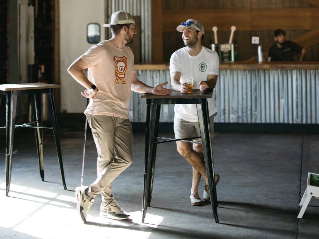 Two men are having a conversation while standing at a high table in a rustic bar. One man is wearing a baseball cap backwards, a white t-shirt, and khaki shorts, and is holding a drink. The other man is wearing a white cap, white t-shirt, and shorts, also holding a drink, and smiling.