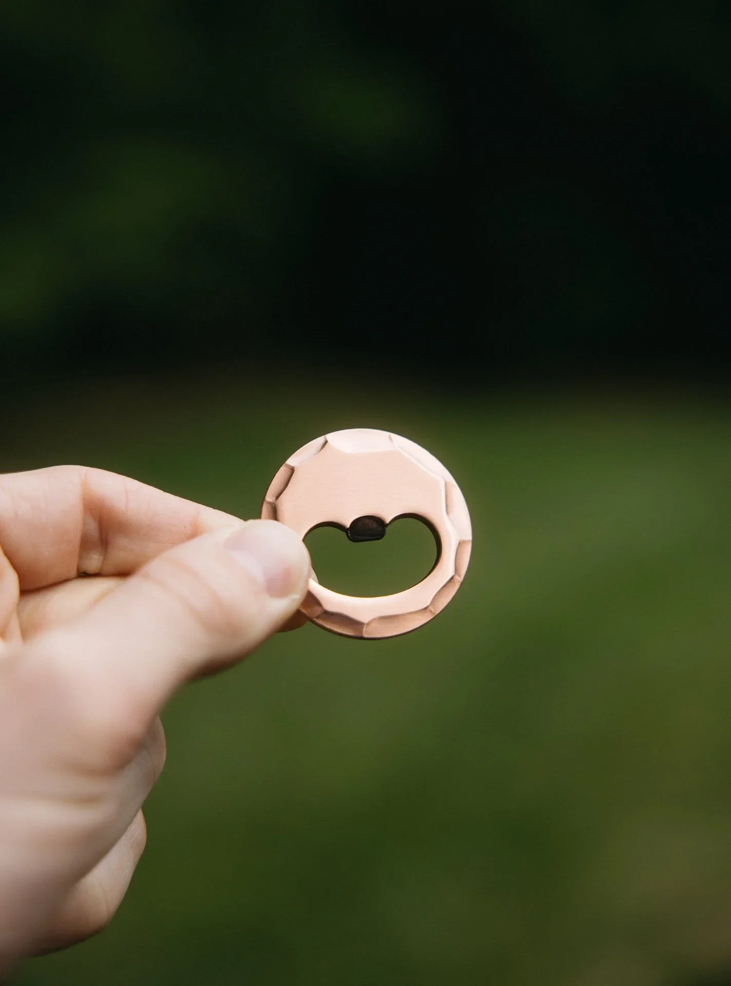 Person holding a shiny copper-colored bottle opener with a circular shape and a cutout in the middle, in front of a blurred green outdoor background.