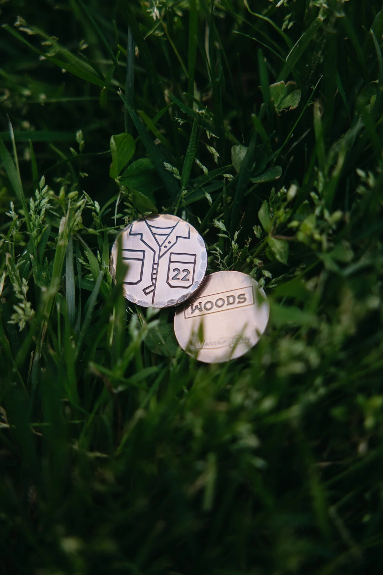 Two wooden tokens with engraved designs, one featuring a shirt with the number 22, and the other with the word "WOODS," placed on green grass.
