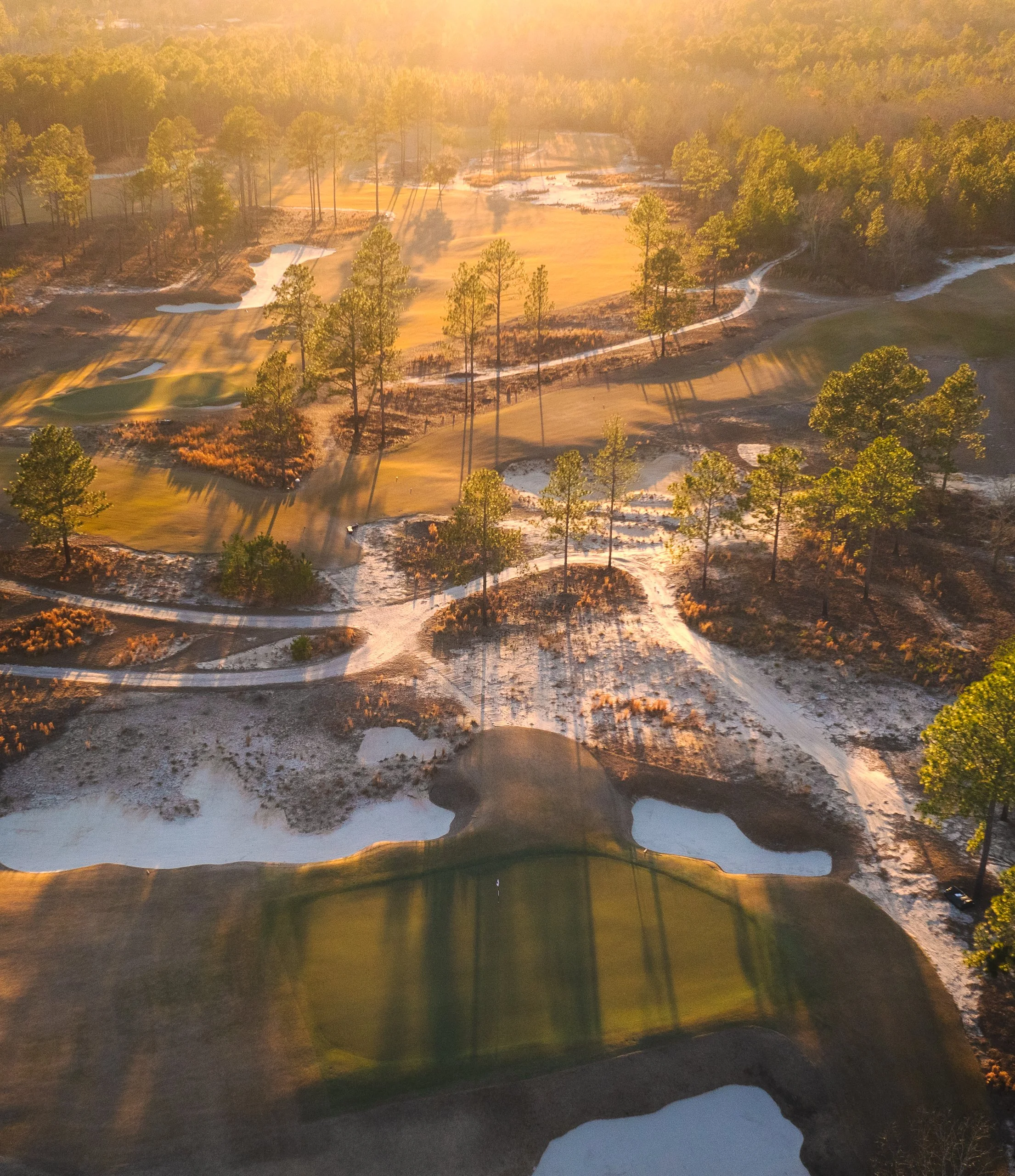 An aerial view of a golf course with sand traps, water hazards, and green fairways amidst trees, under warm sunlight casting long shadows.