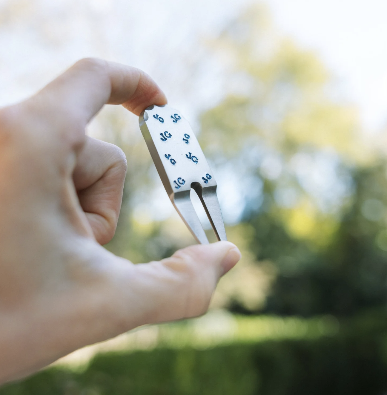 A person holding a small metallic medical tool with a flat, rounded end, outside during daytime with a background of trees and blue sky.